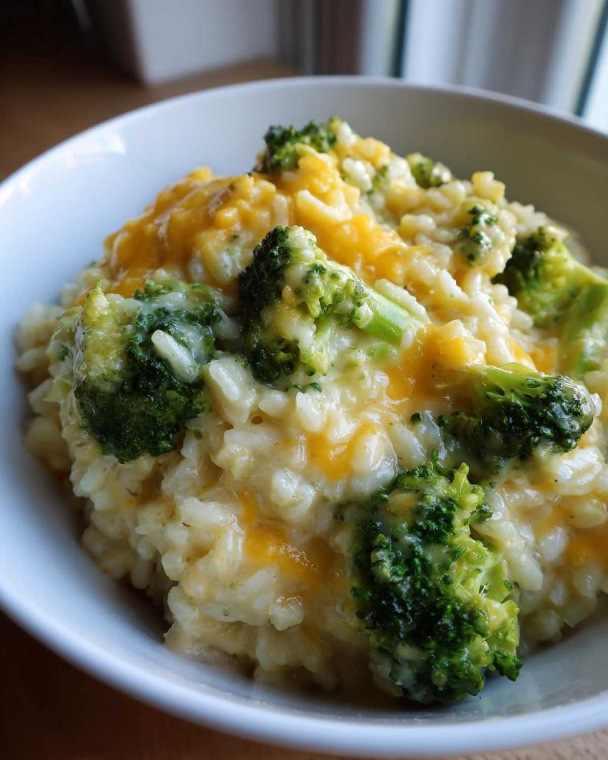 A close-up view of a white bowl filled with One-Pot Creamy Broccoli Cheddar Rice, topped with melted cheddar.