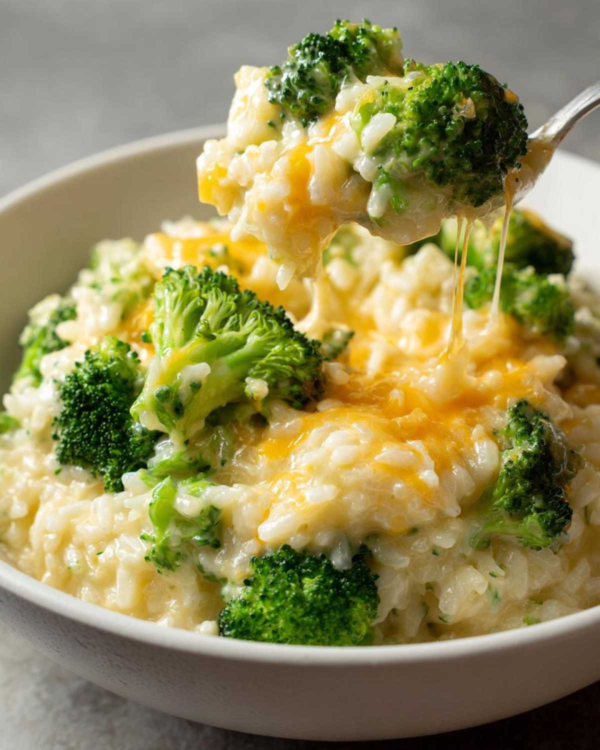 A spoonful of One-Pot Creamy Broccoli Cheddar Rice lifted from a bowl, showing melted cheese stretching between the spoon and the dish.