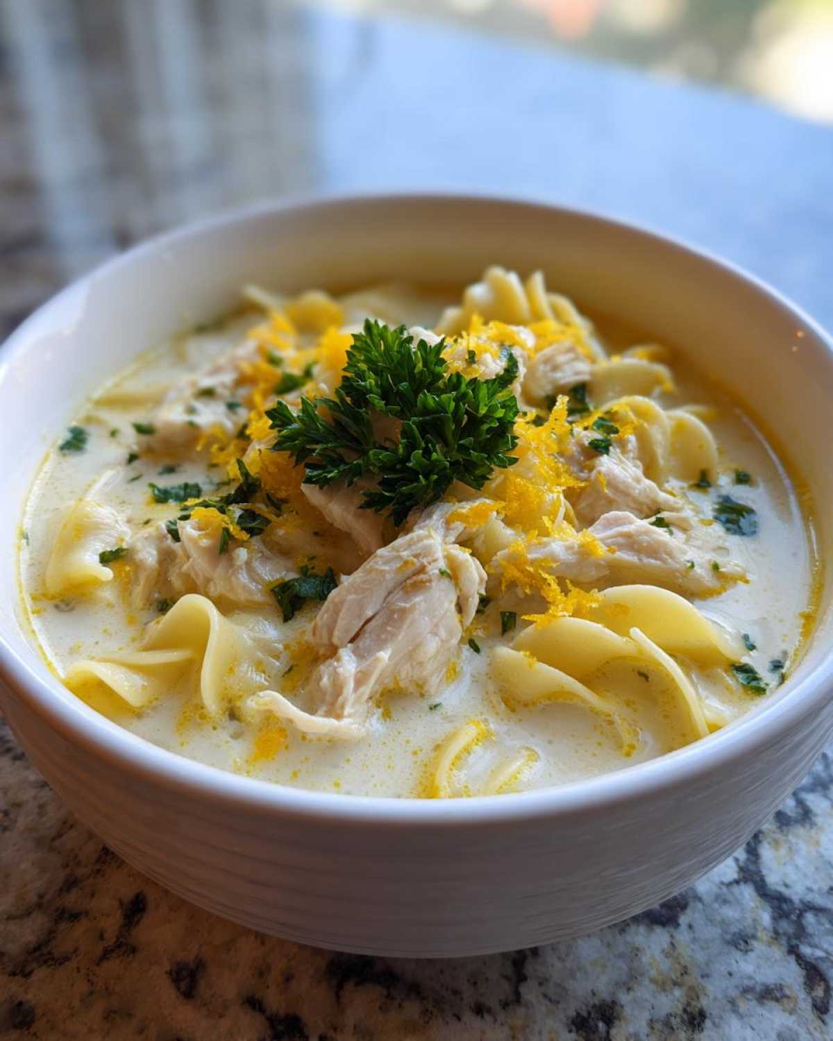 Close-up of a white bowl filled with One-Pot Creamy Lemon Chicken Noodle Soup, topped with shredded chicken, egg noodles, and lemon zest.
