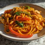 A plate of One-Pot Creamy Thai Red Curry Noodles mixed with chicken, red and green peppers, and fresh basil.