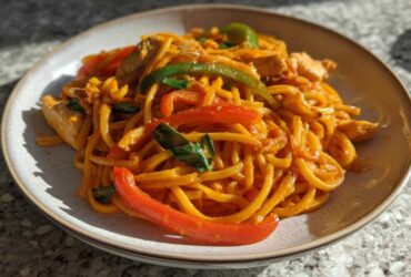A plate of One-Pot Creamy Thai Red Curry Noodles mixed with chicken, red and green peppers, and fresh basil.
