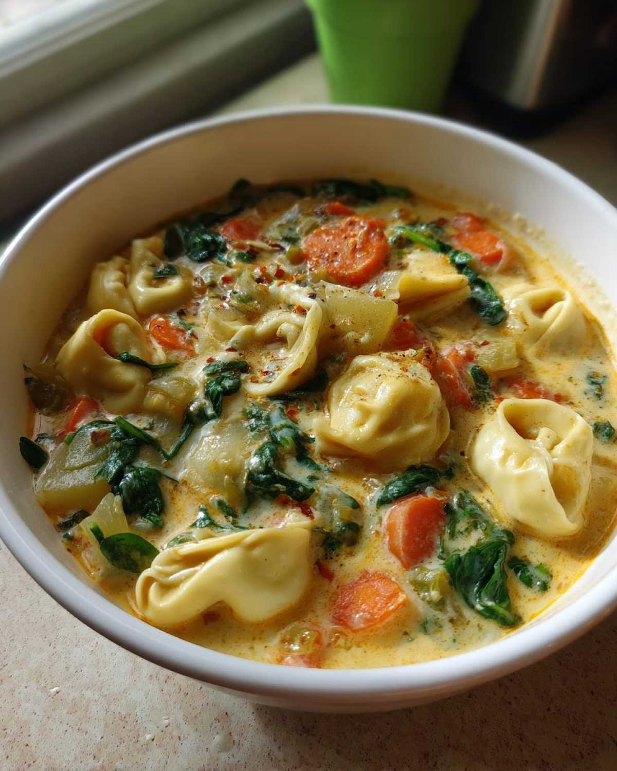 Close-up of a white bowl filled with One-Pot Creamy Tortellini Minestrone soup featuring tortellini, spinach, and carrots.
