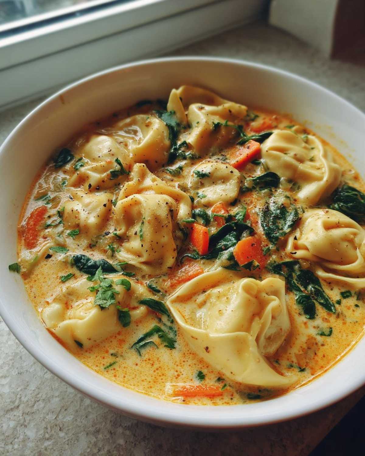 A close-up bowl of One-Pot Creamy Tortellini Minestrone soup with spinach, carrots, and herbs.