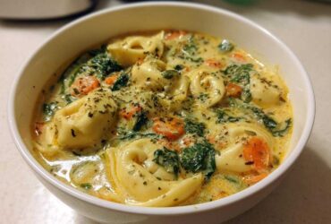 A close-up view of a white bowl filled with One-Pot Creamy Tortellini Minestrone, featuring tortellini, spinach, and carrots in a rich broth.