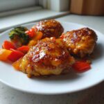 Three pieces of glazed Sheet Pan Ginger Orange Chicken Thighs served with colorful bell peppers and broccoli florets on a white plate.