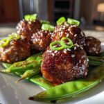 Close-up of glazed Sheet Pan Sticky Sesame Meatballs served over bright green snap peas and garnished with sesame seeds.