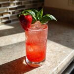 A tall glass of bright pink Sparkling Strawberry Basil Lemonade garnished with a fresh strawberry and basil leaves.