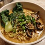 A close-up of a white bowl filled with Vegan Creamy Miso Sesame Noodle Soup, topped with bok choy, mushrooms, and scallions.