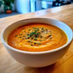 A close-up of a bowl of vibrant orange Vegan Creamy Roasted Carrot Ginger Soup, garnished with fresh chives.