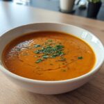A close-up of a bowl of Vegan Creamy Roasted Carrot Ginger Soup, garnished with fresh herbs and a drizzle of oil.