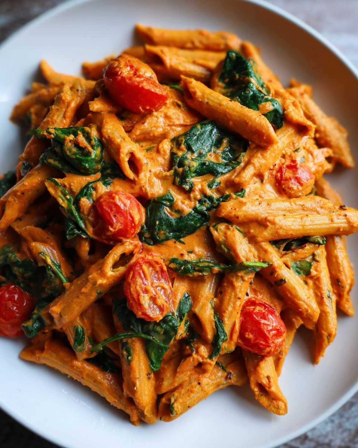 Close-up of Vegan Creamy Roasted Red Pepper Hummus Pasta with spinach and cherry tomatoes in a white bowl.