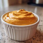 A close-up of thick, vibrant orange Vegan Creamy Sweet Potato Mac Sauce swirled in a white ceramic ramekin.