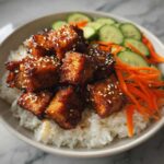 Close-up of a Vegan Crispy Sesame Tempeh Bowl featuring glazed tempeh cubes over white rice with sliced cucumbers and carrots.