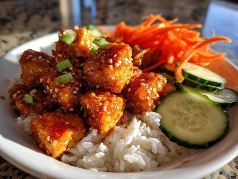 Close-up of a Vegan Crispy Sesame Tempeh Bowl featuring glazed tempeh cubes over rice with cucumber and shredded carrots.