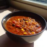 A close-up of a dark bowl filled with rich, red Vegan One-Pot Lentil Taco Soup, featuring visible black beans and corn.
