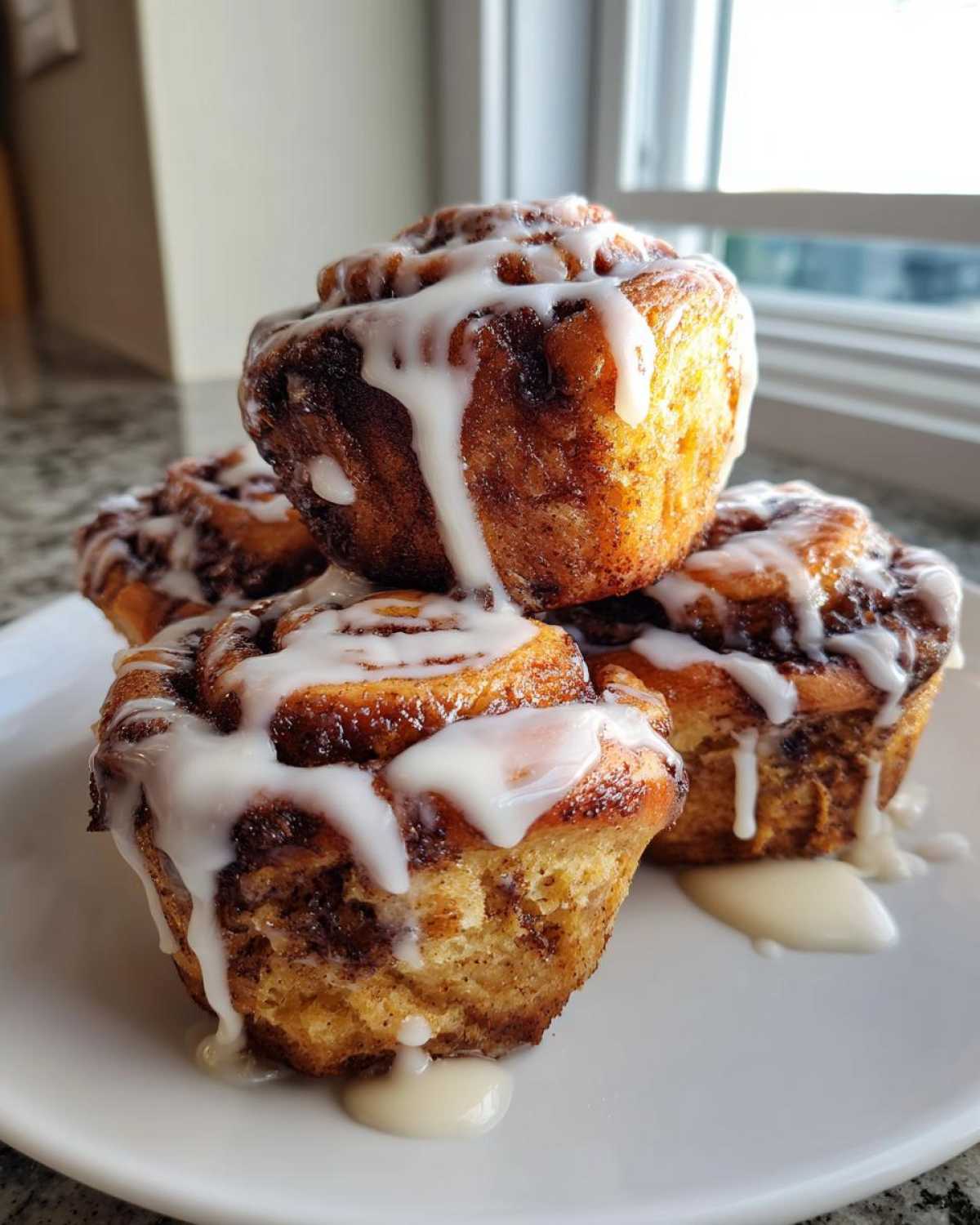 A stack of four moist Banana Bread Batter Cinnamon Buns generously drizzled with white icing, sitting on a white plate.