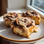 Three freshly baked Banana Bread Cinnamon Rolls covered in thick, dripping glaze sitting on a light plate.