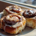 Three freshly baked Banana Bread Cinnamon Rolls covered in thick white icing, sitting on a white plate near a window.