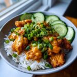 A close-up of a ready-to-eat Bang Bang Chicken Bowl featuring crispy chicken coated in orange sauce over white rice, topped with green onions and sliced cucumbers.