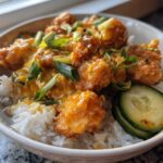 A close-up view of a Bang Bang Chicken Bowl featuring crispy chicken coated in orange sauce over white rice, garnished with scallions and cucumber slices.