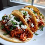 Three assembled Chicken Tinga Tacos Chipotle filled with shredded chicken in red sauce, topped with cotija cheese and cilantro.