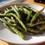 A close-up of glossy, roasted Crack Green Beans seasoned with garlic bits, served on a white plate.