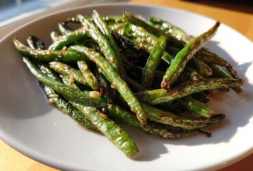 A close-up of glossy, roasted Crack Green Beans seasoned with garlic bits, served on a white plate.