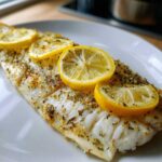 Close-up of a flaky, white Easy Baked Fish fillet seasoned with herbs and topped with bright lemon slices.