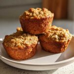 Three golden brown Oat Muffins Breakfast stacked on a light plate, topped with visible rolled oats.