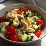 Close-up of a bowl containing a high protein dinner idea: grilled chicken, quinoa, cherry tomatoes, cucumber, and feta cheese.
