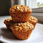 Three golden brown Breakfast Muffins Peanut Free stacked on a white plate, topped with rolled oats.