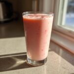 A tall glass filled with pink, frothy Strawberry Brazilian Lemonade sitting on a sunlit countertop.