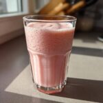A glass filled with pink, frothy Strawberry Brazilian Lemonade sitting on a kitchen counter in bright sunlight.