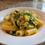 A close-up of a white bowl filled with Tasty Lemon Zucchini Rigatoni Pasta, featuring yellow sauce, green zucchini chunks, and basil.