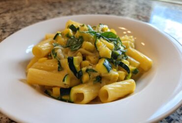 A close-up of a white bowl filled with Tasty Lemon Zucchini Rigatoni Pasta, featuring yellow sauce, green zucchini chunks, and basil.